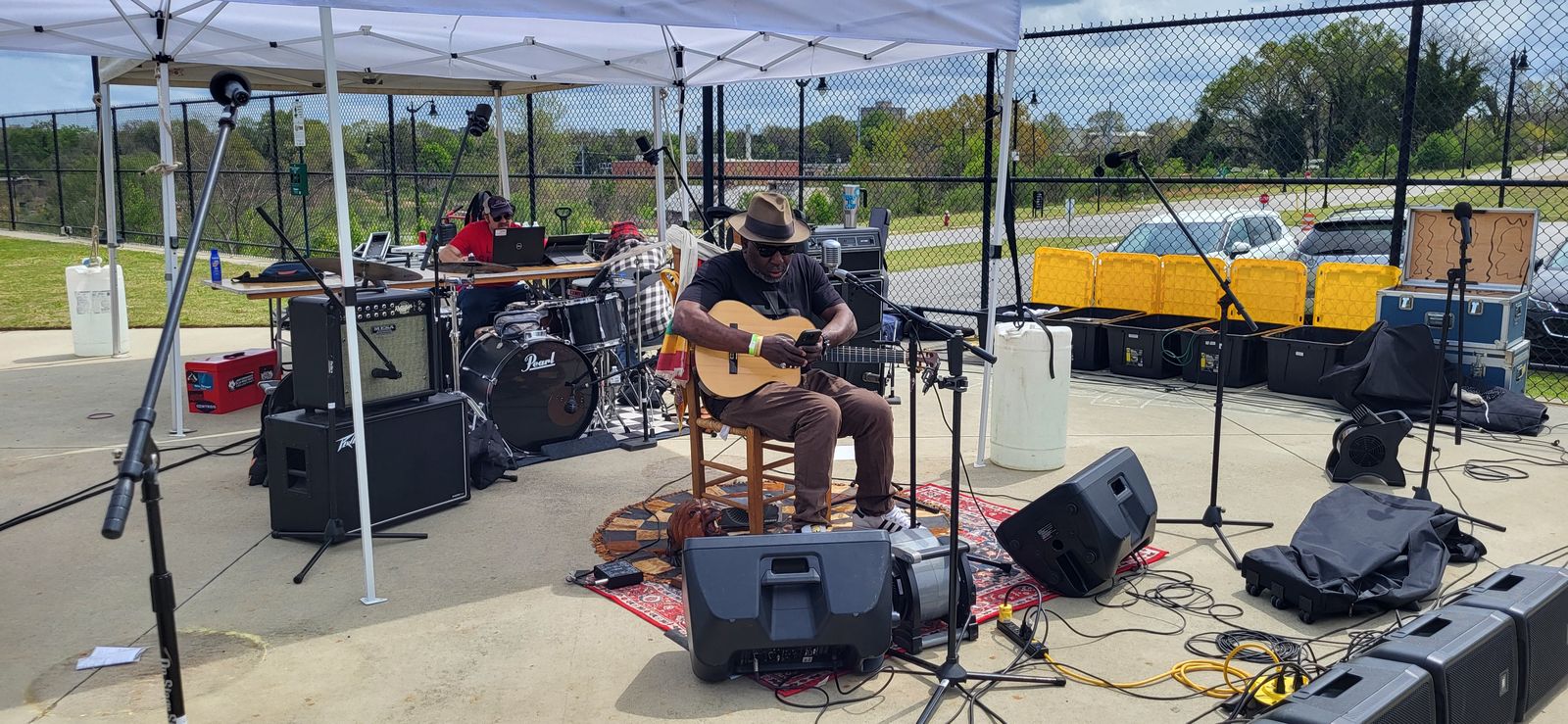 An outdoor stage under a white canopy, with a seated guitarist, a drum kit and speakers on a rug.