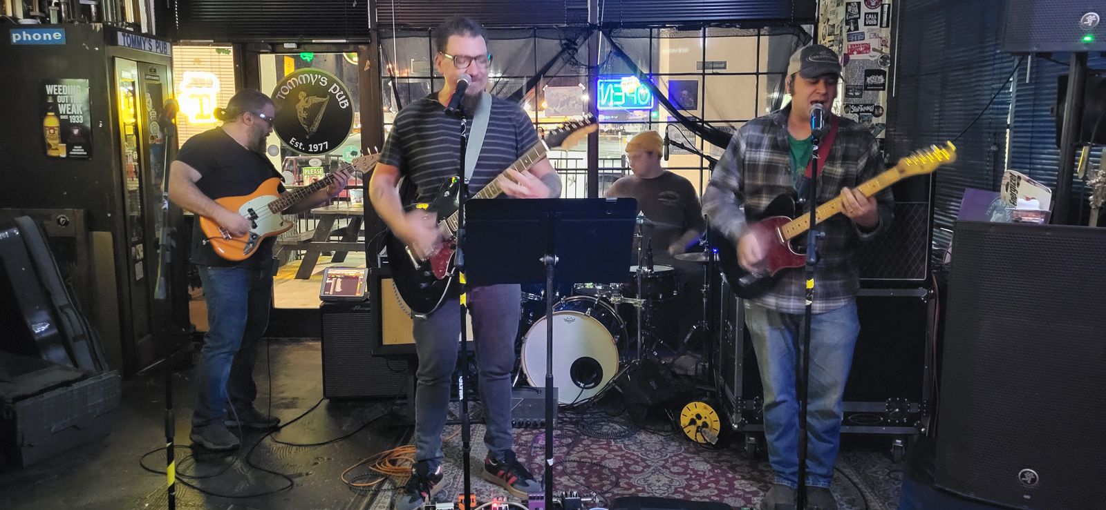 A four-piece rock band plays on a small pub stage, with three guitarists out front and a drummer behind them.