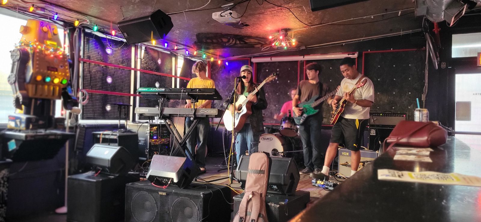 A band plays at Art Bar with a singer on acoustic guitar, an electric guitarist, a bassist and a keyboardist under string lights.