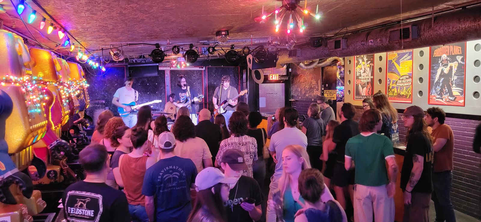 A packed crowd faces a band in a colorful bar with string lights, neon art and gig posters on the walls.