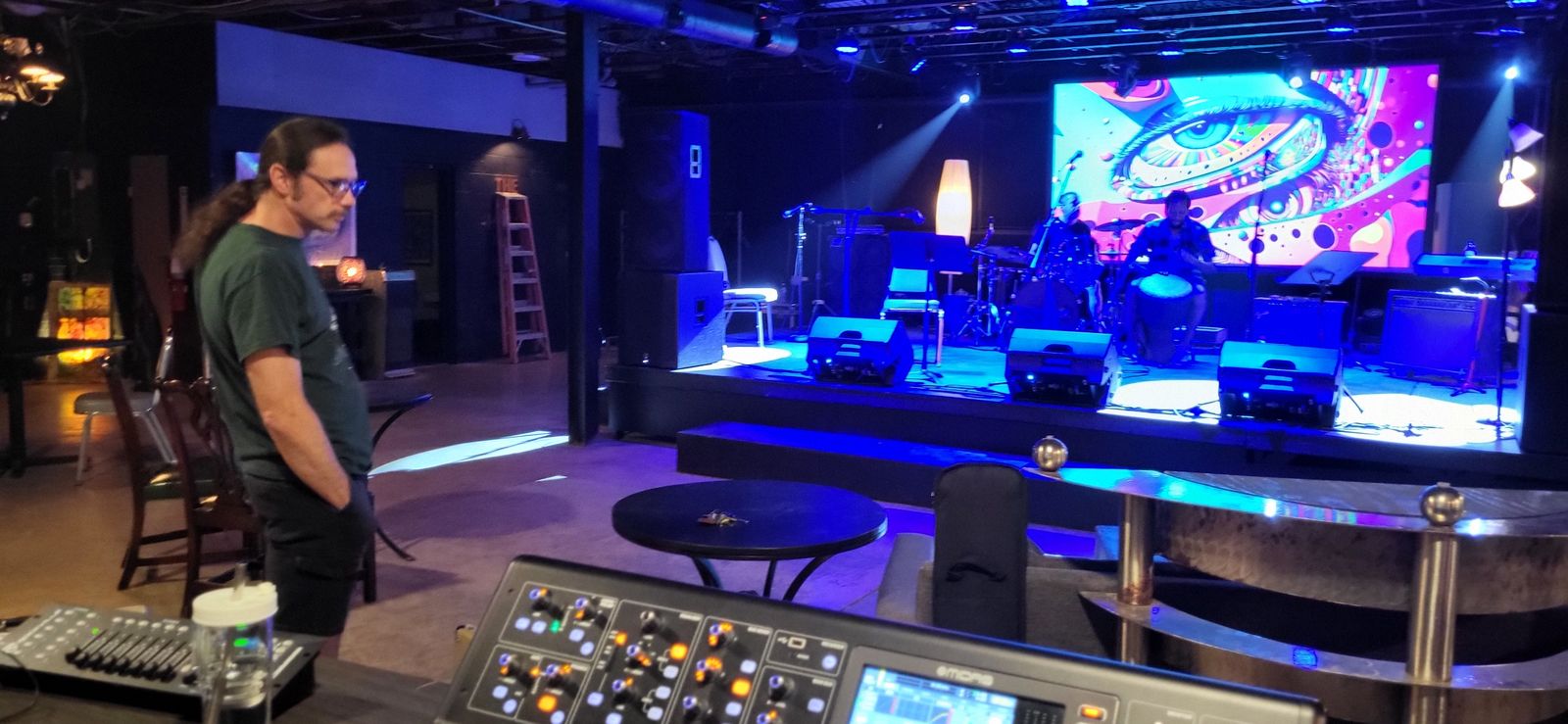 A big stage washed in blue light with a psychedelic eye graphic on the video wall. A mixing board sits in the foreground.