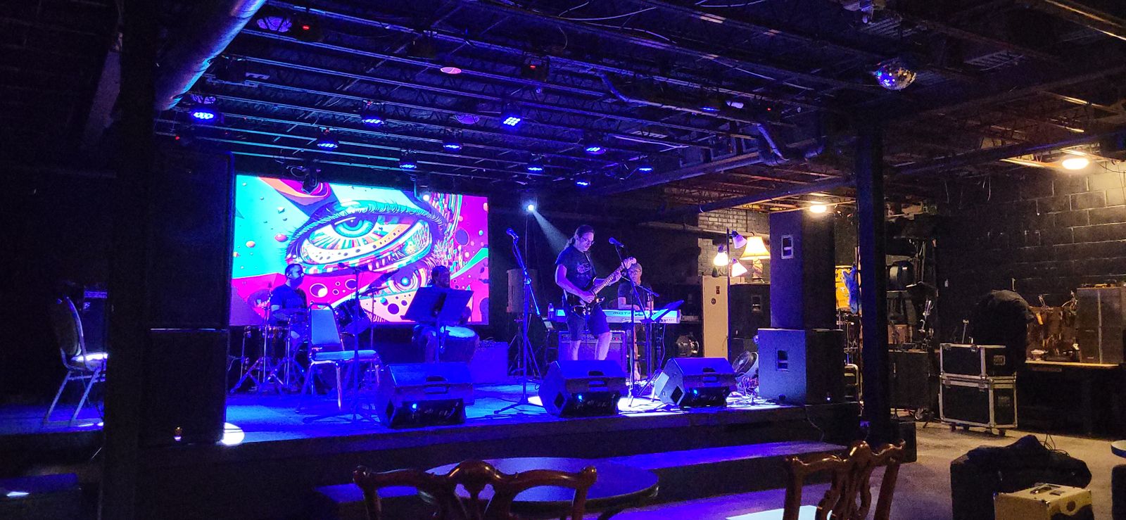 A wide shot of a warehouse venue with musicians performing on a blue-lit stage under exposed ceiling beams.