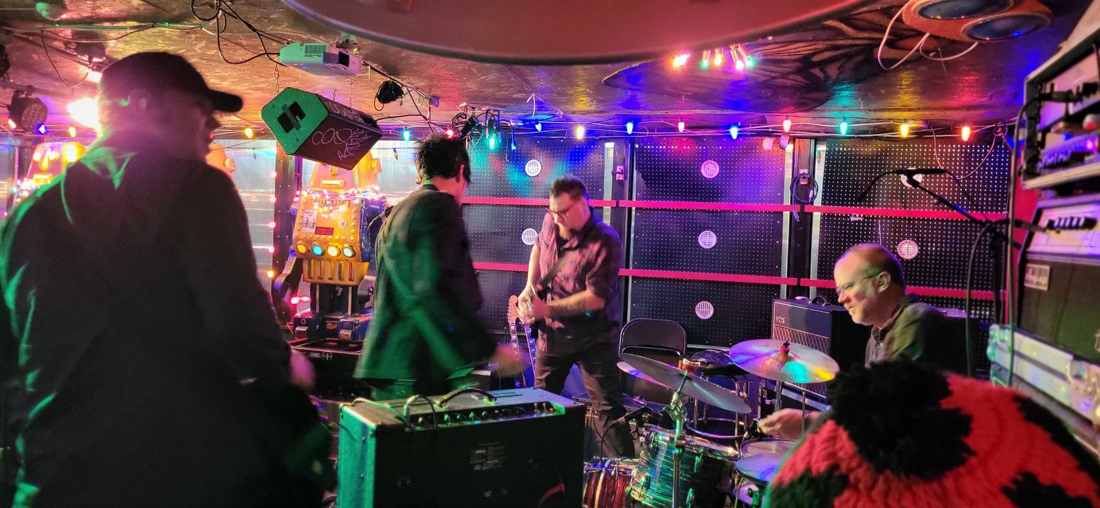 The view from the crowd toward a neon-lit stage, with musicians and gear surrounded by fans.
