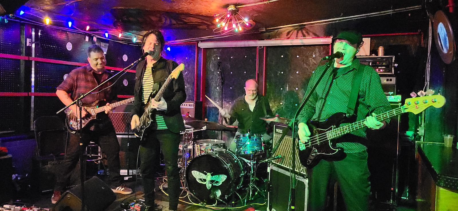 A band on a small stage lit in red, purple and green, with string lights overhead. Two guitarists, a bassist in green and a drummer.