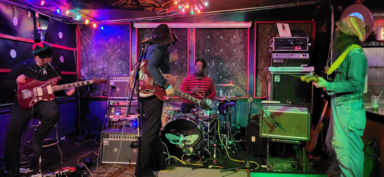 A late-night band on a colorfully lit stage, with guitarists, a drummer and amps along the wall.